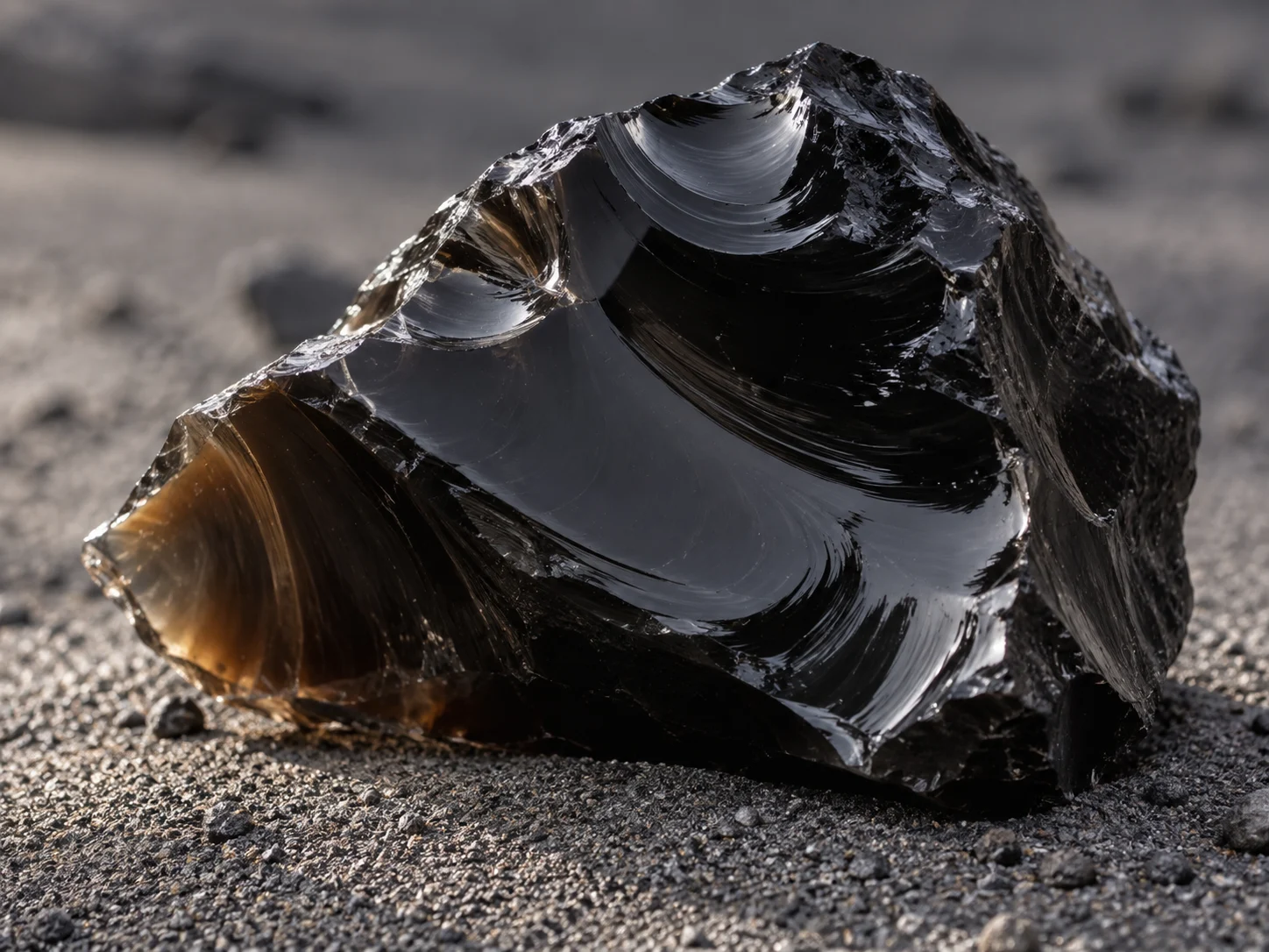 A glossy black obsidian stone with smooth curved fractures on gray volcanic sand