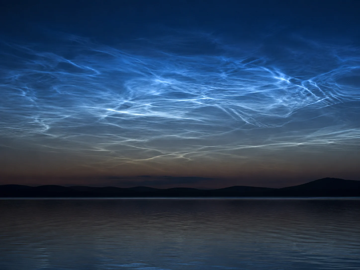 Silver-blue noctilucent cloud ripples glowing above a dark lake after sunset