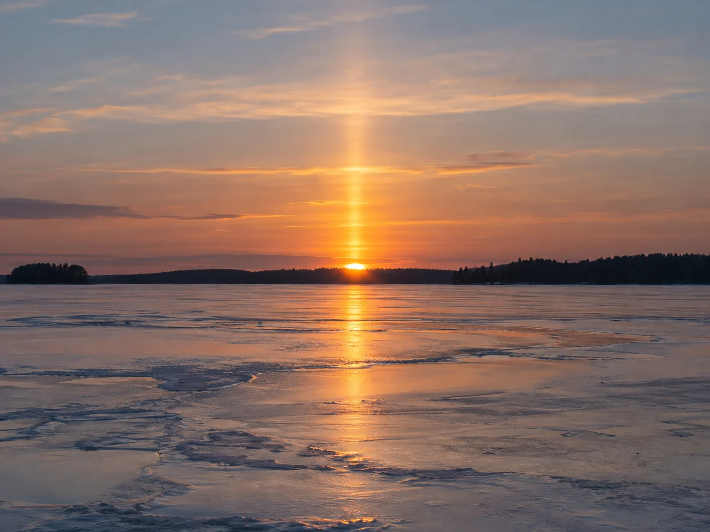 A vertical golden sun pillar above a low sunset over a frozen lake