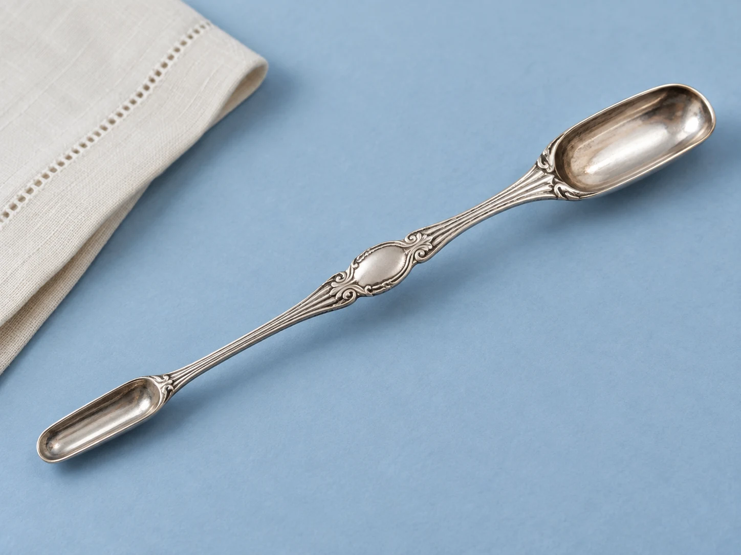 A long double-ended silver marrow scoop with one narrow end and one wider end on a blue surface